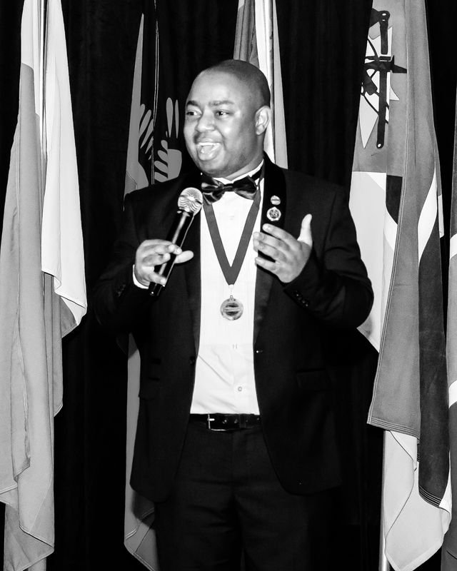 Frank Tsuro in black tie, speaking into a handheld microphone in front of a row of flags. Black and white.