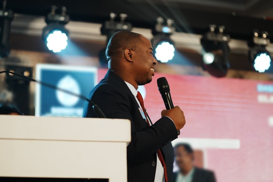 Frank Tsuro speaking at a lectern with a handheld microphone. Wide frame in colour.