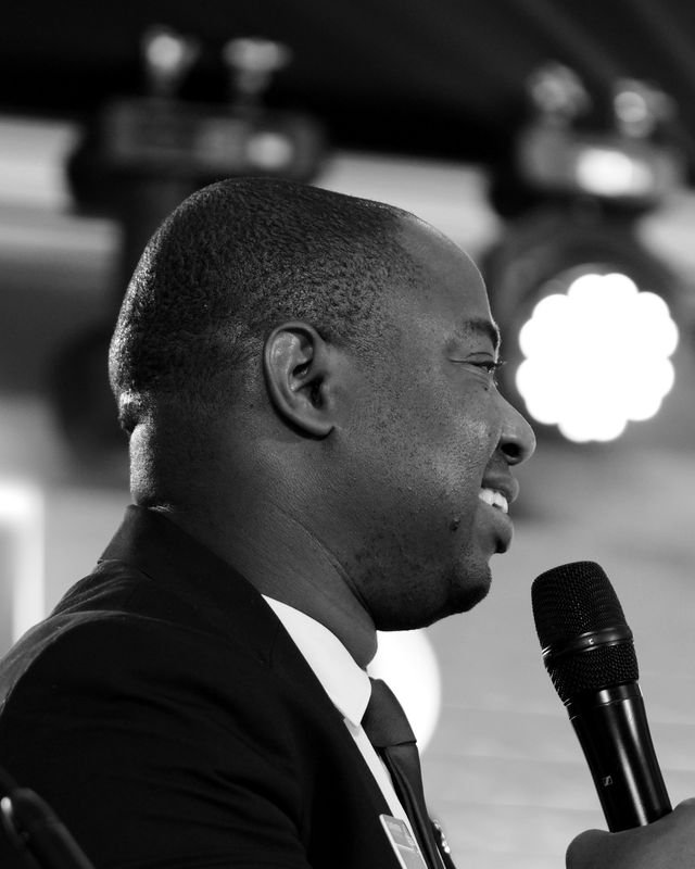Frank Tsuro in profile, smiling at a microphone under stage lights. Black and white.